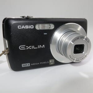 Puede incluir: Cámara digital Casio Exilim negra con lente plateada. La cámara tiene una resolución de 12,1 megapíxeles y un zoom óptico de 3x. La cámara está sobre una superficie blanca.