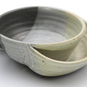 May include: A ceramic bowl with a single handle, featuring a two-tone glaze design. One side of the bowl is a dark charcoal gray, while the other side is a light cream color with subtle brown speckles. The surface has visible horizontal ridges.