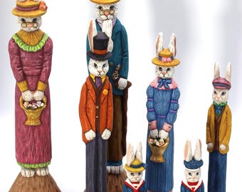 May include: A collection of seven hand-painted wooden rabbit figurines standing on textured brown bases. The rabbits are dressed in colourful Victorian-style clothing, including hats, jackets, and dresses, with some holding small baskets or accessories.