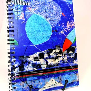 May include: A spiral-bound notebook featuring a vibrant blue cover with abstract white circular line art, black geometric shapes, and accents of red and green. The cover design includes layered textures and a prominent light blue oval shape.