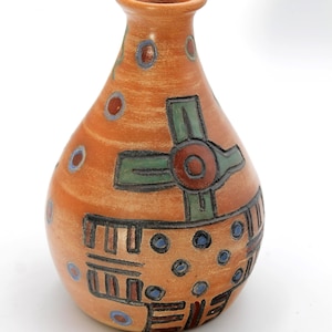 May include: A terracotta-colored ceramic vase with a narrow neck and a rounded body. The surface features hand-painted geometric patterns in green, black, and blue, including circular dots, cross shapes, and linear motifs across the exterior.