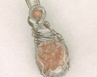 May include: A handcrafted pendant featuring a druzy gemstone in a teardrop shape, encased in a silver wire wrap. The pendant has a light peach color and a small, round, peach-colored bead at the top.