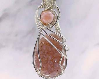 May include: A handcrafted pendant featuring a teardrop-shaped, pinkish-brown stone wrapped in silver wire. A smaller, round, pink stone is set above the larger stone. The pendant hangs from a silver chain, creating a unique jewelry piece.