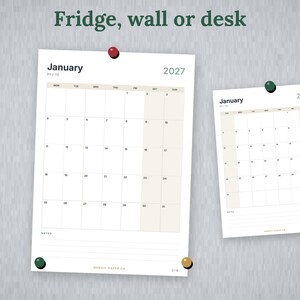 May include: Two white paper calendars for January 2027, each featuring a grid layout with beige columns for weekends. The larger calendar is secured by a red pin and a green pin, while the smaller one is held by a green pin. Text reads Fridge, wall or desk.
