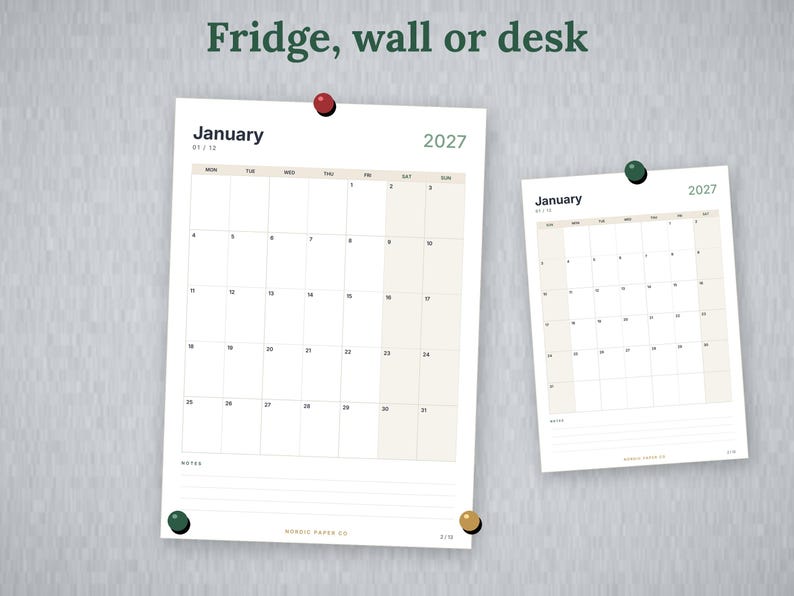 May include: Two white paper calendars for January 2027, each featuring a grid layout with beige columns for weekends. The larger calendar is secured by a red pin and a green pin, while the smaller one is held by a green pin. Text reads Fridge, wall or desk.