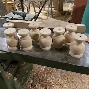 May include: A set of eight unfinished light-coloured wooden furniture legs, each featuring a rounded, bulbous midsection and a flat circular top with a small centred hole, arranged on a flat metal surface in a workshop setting.