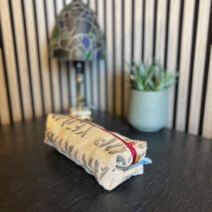 Könnte beinhalten: Eine braune Stiftetui aus Jute mit einem roten Reißverschluss. Das Etui liegt auf einer schwarzen Oberfläche.