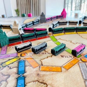 Könnte beinhalten: Ein Spielbrett mit einer Karte von Europa und Zugwagen in verschiedenen Farben, darunter grün, rosa und grau. Die Zugwagen befinden sich auf dem Brett, und der Text "FR", "MUNCHEN", "WIEN" ist sichtbar.