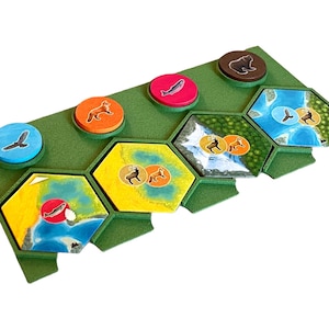 Può includere: Un tabellone da gioco verde con tessere esagonali che presentano varie illustrazioni di animali e natura. Le tessere sono disposte in fila e includono immagini di una volpe, un cervo, un pesce, un orso e un uccello.