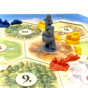 Puede incluir: Una figurita de plástico gris de una persona que lleva una pila de cuatro ovejas grises en un tablero de juego con un paisaje marrón, amarillo y rojo. El tablero tiene los números 9 y 10.