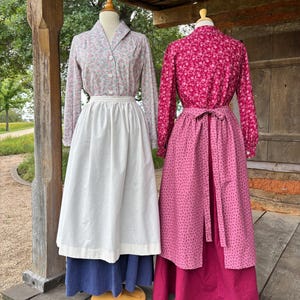 以下が含まれることがあります： マネキンに展示された2つの衣装。左の衣装は、明るい花柄の長袖シャツ、襟とボタン、無地の白いエプロン、無地の青いスカートで構成されています。右の衣装は、濃いピンクの花柄シャツと、無地の濃いピンクのスカートの上に合わせた同柄のエプロンで構成されています。