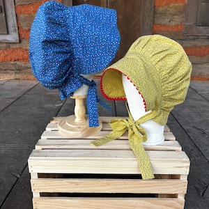 Puede incluir: Dos gorros de tela sobre soportes. El gorro de la izquierda es azul con un pequeño estampado floral multicolor y lazos laterales. El gorro de la derecha es amarillo con un fino estampado floral negro, un borde festoneado rojo y lazos laterales.