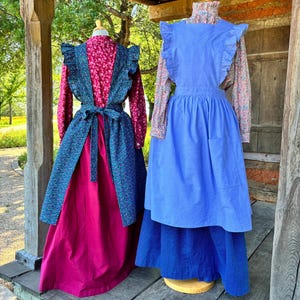 May include: Two mannequins wearing long-sleeved floral blouses under solid-coloured pinafore aprons with ruffled shoulder straps. One apron is dark blue with a small floral print, worn over a magenta skirt. The other is solid blue over a blue skirt.