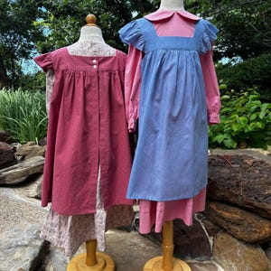 May include: Two dresses displayed on mannequins. The left dress is a dark pink pinafore with a white dot pattern over a floral print dress. The right dress is a blue pinafore with ruffled shoulders worn over a solid pink long-sleeved dress.