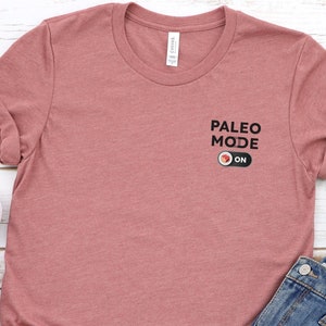 Puede incluir: Una camiseta de color malva jaspeado con el texto "Paleo Mode ON" y un gráfico de interruptor de palanca rojo.