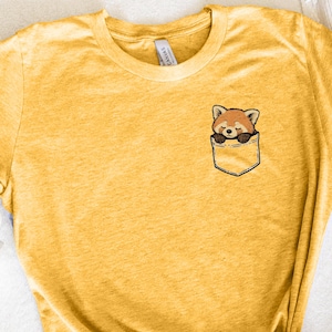 May include: Mustard yellow t-shirt featuring a pocket with a sleeping red panda graphic. The red panda has brown and white fur and is nestled in the pocket. The shirt is made of a soft material.