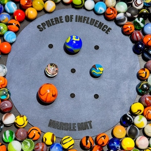 Op de afbeelding: Een zwarte ronde marmermat met de tekst "Sphere of Influence" en "Marble Mat" erop gedrukt. De mat is omringd door een verscheidenheid aan kleurrijke marmers.