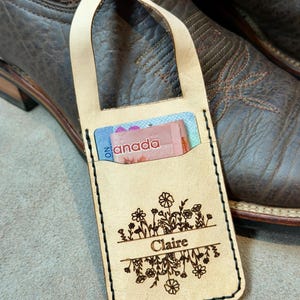 May include: A light tan leather luggage tag with a loop and brass stud closure. The front features a laser-engraved floral design with the name Claire in the centre. A Canadian banknote and a card are tucked into the pocket of the tag.