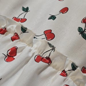 K&ouml;nnte beinhalten: Nahaufnahme eines wei&szlig;en Stoffes mit einem sich wiederholenden Muster aus roten Kirschen und gr&uuml;nen Bl&auml;ttern. Der Stoff hat einen ger&uuml;schten Rand. Die Kirschen sind stilisiert mit schwarzen Stielen und sind &uuml;ber das Material verstreut.