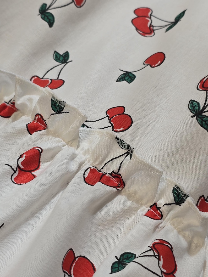 K&ouml;nnte beinhalten: Nahaufnahme eines wei&szlig;en Stoffes mit einem sich wiederholenden Muster aus roten Kirschen und gr&uuml;nen Bl&auml;ttern. Der Stoff hat einen ger&uuml;schten Rand. Die Kirschen sind stilisiert mit schwarzen Stielen und sind &uuml;ber das Material verstreut.