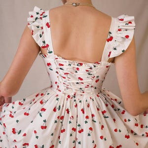 K&ouml;nnte beinhalten: Wei&szlig;es Sommerkleid mit rotem Kirschmuster. Das Kleid hat ger&uuml;schte Fl&uuml;gel&auml;rmel, einen Korsettr&uuml;cken und einen weiten Rock. Die Kirschen haben gr&uuml;ne Stiele und Bl&auml;tter. Ein klassisches, feminines Sommerkleid.