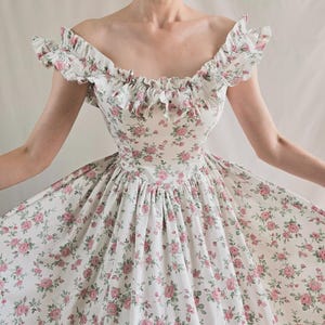 May include: A white dress featuring a repeating pattern of small pink roses and green leaves. The garment has an off-the-shoulder neckline with ruffled trim along the collar and a fitted bodice that transitions into a full, flared skirt.