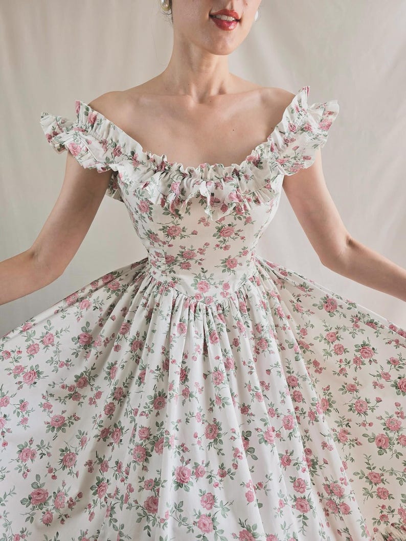 May include: A white dress featuring a repeating pattern of small pink roses and green leaves. The garment has an off-the-shoulder neckline with ruffled trim along the collar and a fitted bodice that transitions into a full, flared skirt.