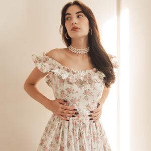 May include: A white off-the-shoulder dress featuring a delicate pink and green floral pattern, a ruffled neckline, and a fitted bodice. The garment is accessorized with a multi-strand pearl choker necklace and gold hoop earrings.