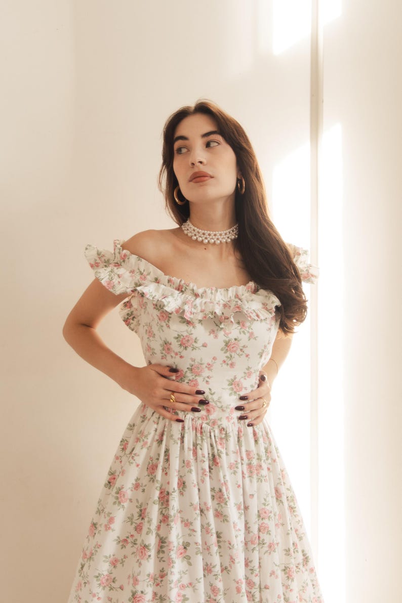 May include: A white off-the-shoulder dress featuring a delicate pink and green floral pattern, a ruffled neckline, and a fitted bodice. The garment is accessorized with a multi-strand pearl choker necklace and gold hoop earrings.