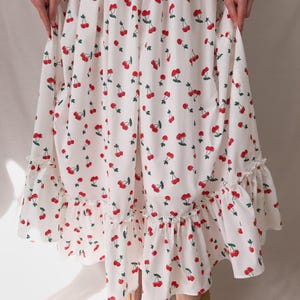 K&ouml;nnte beinhalten: Wei&szlig;es Kleid mit Kirschmuster. Das Kleid hat ein sich wiederholendes Muster aus roten Kirschen mit gr&uuml;nen Bl&auml;ttern auf dem Stoff. Das Kleid hat einen R&uuml;schensaum und eine flie&szlig;ende Silhouette.