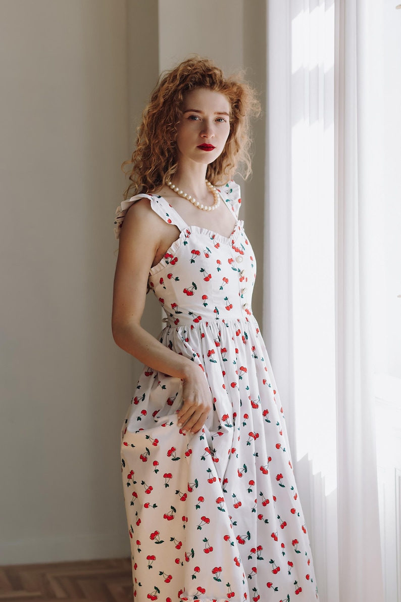 K&ouml;nnte beinhalten: Ein wei&szlig;es Sommerkleid mit rotem Kirschmuster. Das Kleid hat ein figurbetontes Oberteil, R&uuml;schentr&auml;ger und einen flie&szlig;enden Rock. Das Model tr&auml;gt eine Perlenkette. Das Kleid ist im Vintage-Stil gehalten.