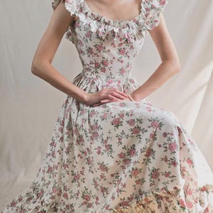 May include: A floor-length dress featuring a white base with a pink and green floral rose pattern. The garment includes a scoop neckline with ruffled trim along the shoulders and bodice, a fitted waist, and a wide ruffled hem at the bottom edge.
