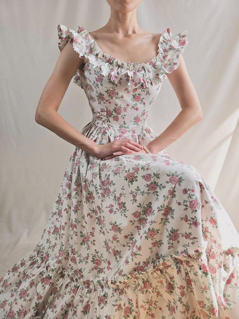 May include: A floor-length dress featuring a white base with a pink and green floral rose pattern. The garment includes a scoop neckline with ruffled trim along the shoulders and bodice, a fitted waist, and a wide ruffled hem at the bottom edge.