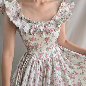 May include: A white dress featuring a repeating pattern of small pink roses and green leaves. The garment has a wide scoop neckline adorned with a ruffled trim that extends over the shoulders, a fitted bodice, and a gathered skirt with a full silhouette.