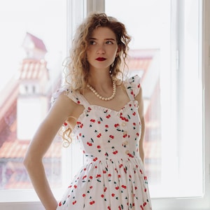 K&ouml;nnte beinhalten: Wei&szlig;es Sommerkleid mit rotem Kirschmuster. Das Kleid hat einen quadratischen Ausschnitt, eine Knopfleiste vorne und R&uuml;schentr&auml;ger. Eine Perlenkette sorgt f&uuml;r Eleganz. Ideal f&uuml;r einen Sommertag.