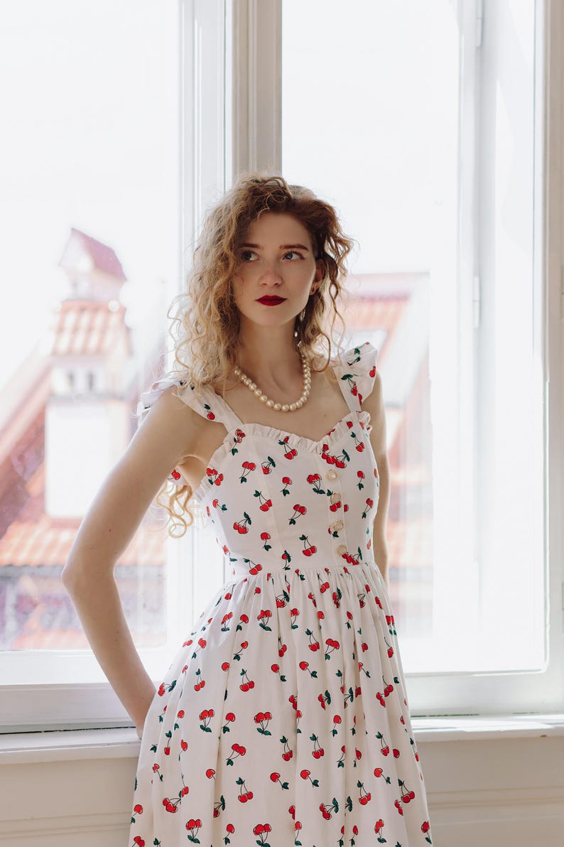 K&ouml;nnte beinhalten: Wei&szlig;es Sommerkleid mit rotem Kirschmuster. Das Kleid hat einen quadratischen Ausschnitt, eine Knopfleiste vorne und R&uuml;schentr&auml;ger. Eine Perlenkette sorgt f&uuml;r Eleganz. Ideal f&uuml;r einen Sommertag.
