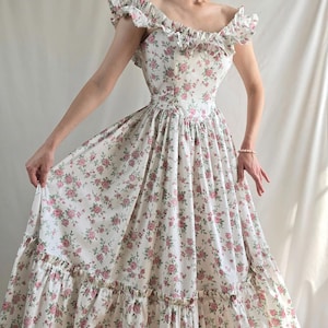 May include: A floor-length dress featuring an off-the-shoulder neckline with ruffled trim and a fitted bodice. The fabric is white with a repeating pattern of small pink roses and green leaves. The skirt is full with a wide ruffled hem at the bottom.