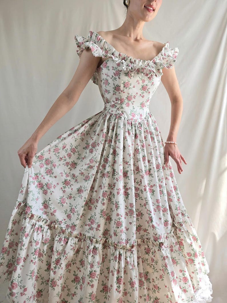 May include: A floor-length dress featuring an off-the-shoulder neckline with ruffled trim and a fitted bodice. The fabric is white with a repeating pattern of small pink roses and green leaves. The skirt is full with a wide ruffled hem at the bottom.