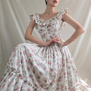 May include: A floor-length dress featuring a white base with a repeating pink rose and green leaf floral pattern. The garment includes a square neckline, ruffled cap sleeves, a fitted bodice, and a tiered skirt with a ruffled hemline detail.
