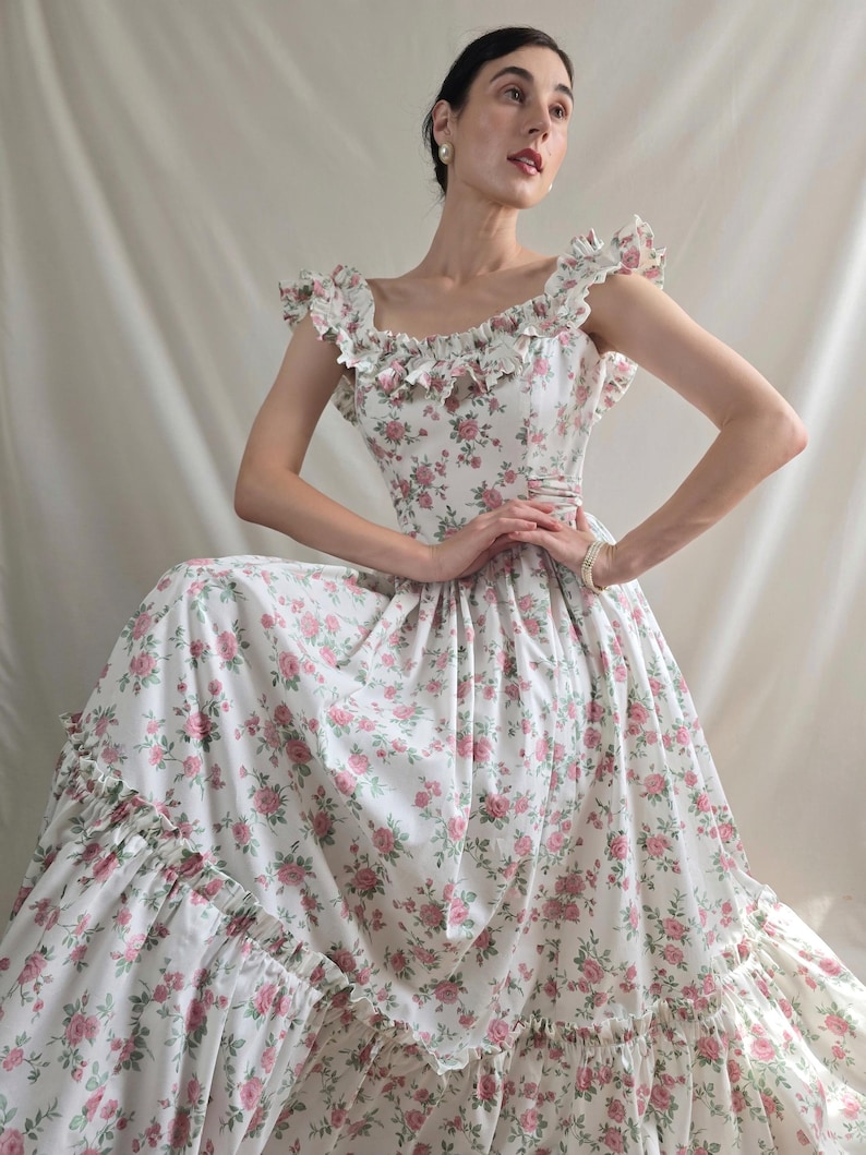 May include: A floor-length dress featuring a white base with a repeating pink rose and green leaf floral pattern. The garment includes a square neckline, ruffled cap sleeves, a fitted bodice, and a tiered skirt with a ruffled hemline detail.