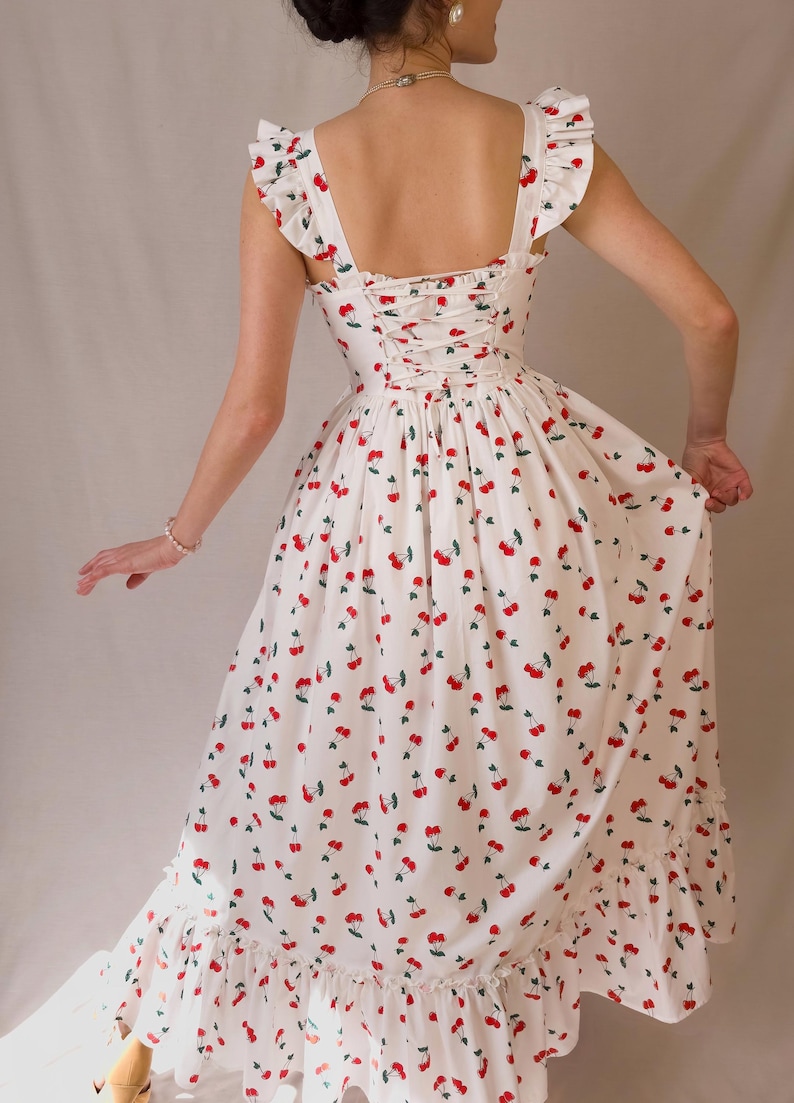 K&ouml;nnte beinhalten: Ein wei&szlig;es Kleid mit Kirschmuster. Das Kleid hat einen quadratischen Ausschnitt, R&uuml;schen&auml;rmel und einen R&uuml;cken im Korsett-Stil. Der Rock hat einen R&uuml;schensaum. Die Kirschen sind rot mit gr&uuml;nen Bl&auml;ttern.