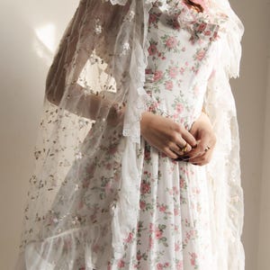 May include: A white dress featuring a pink and green floral pattern with a ruffled neckline and a pink ribbon tie. A sheer white lace shawl with intricate floral embroidery and ruffled edges is draped over the shoulders and head.