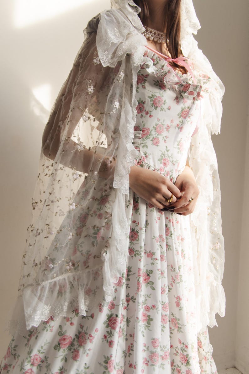 May include: A white dress featuring a pink and green floral pattern with a ruffled neckline and a pink ribbon tie. A sheer white lace shawl with intricate floral embroidery and ruffled edges is draped over the shoulders and head.