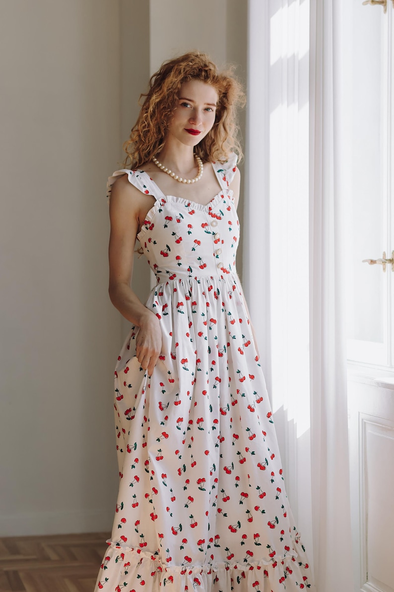 K&ouml;nnte beinhalten: Ein wei&szlig;es Sommerkleid mit rotem Kirschmuster. Das Kleid hat R&uuml;schentr&auml;ger, ein figurbetontes Oberteil und einen flie&szlig;enden Rock. Das Kleid ist mit kleinen roten Kirschillustrationen verziert und hat einen R&uuml;schensaum. Eine Perlenkette ist ebenfalls sichtbar.