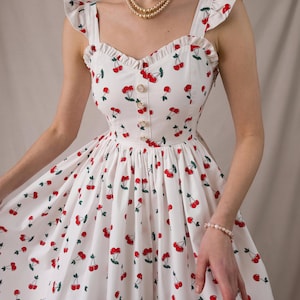 K&ouml;nnte beinhalten: Wei&szlig;es Sommerkleid mit rotem Kirschmuster. Das Kleid hat ger&uuml;schte Tr&auml;ger, ein figurbetontes Oberteil mit Kn&ouml;pfen und einen ausgestellten Rock. Eine Perlenkette und ein Armband sind ebenfalls zu sehen.