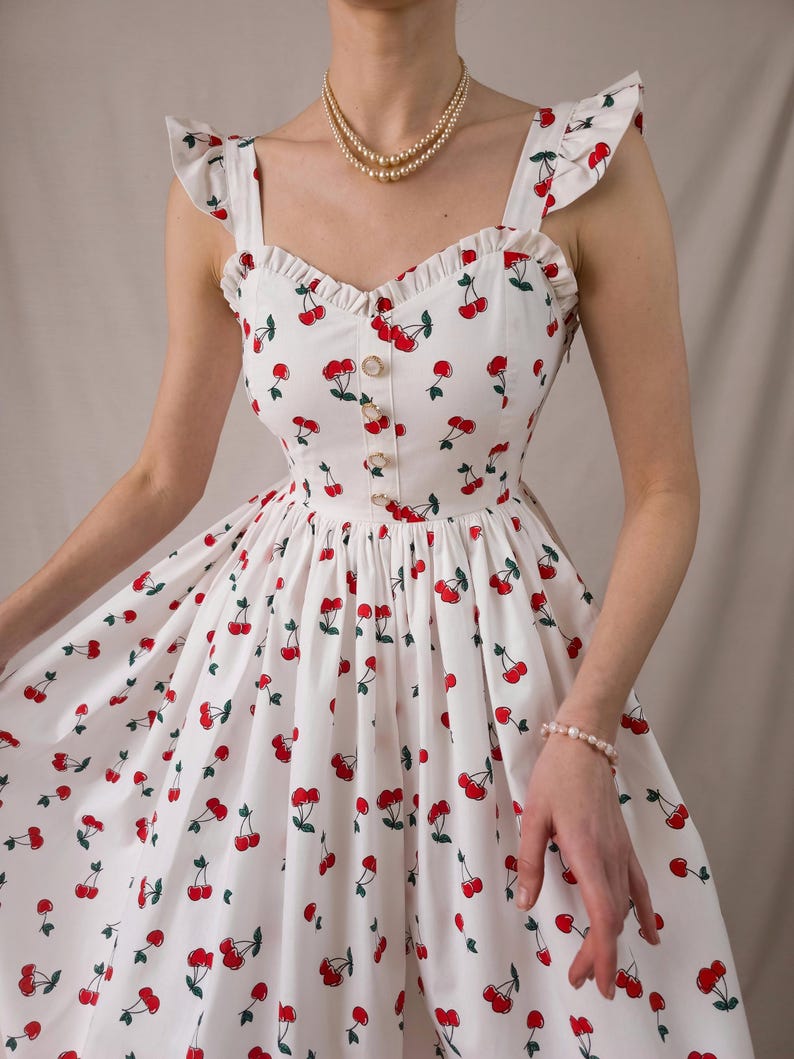 K&ouml;nnte beinhalten: Wei&szlig;es Sommerkleid mit rotem Kirschmuster. Das Kleid hat ger&uuml;schte Tr&auml;ger, ein figurbetontes Oberteil mit Kn&ouml;pfen und einen ausgestellten Rock. Eine Perlenkette und ein Armband sind ebenfalls zu sehen.