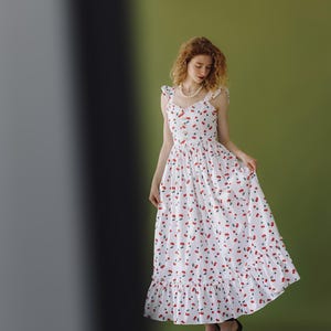 K&ouml;nnte beinhalten: Ein wei&szlig;es Maxikleid mit rotem Kirschmuster. Das &auml;rmellose Kleid hat einen R&uuml;schensaum und Tr&auml;ger. Die Person tr&auml;gt schwarze Abs&auml;tze. Der Hintergrund ist einfarbig gr&uuml;n.