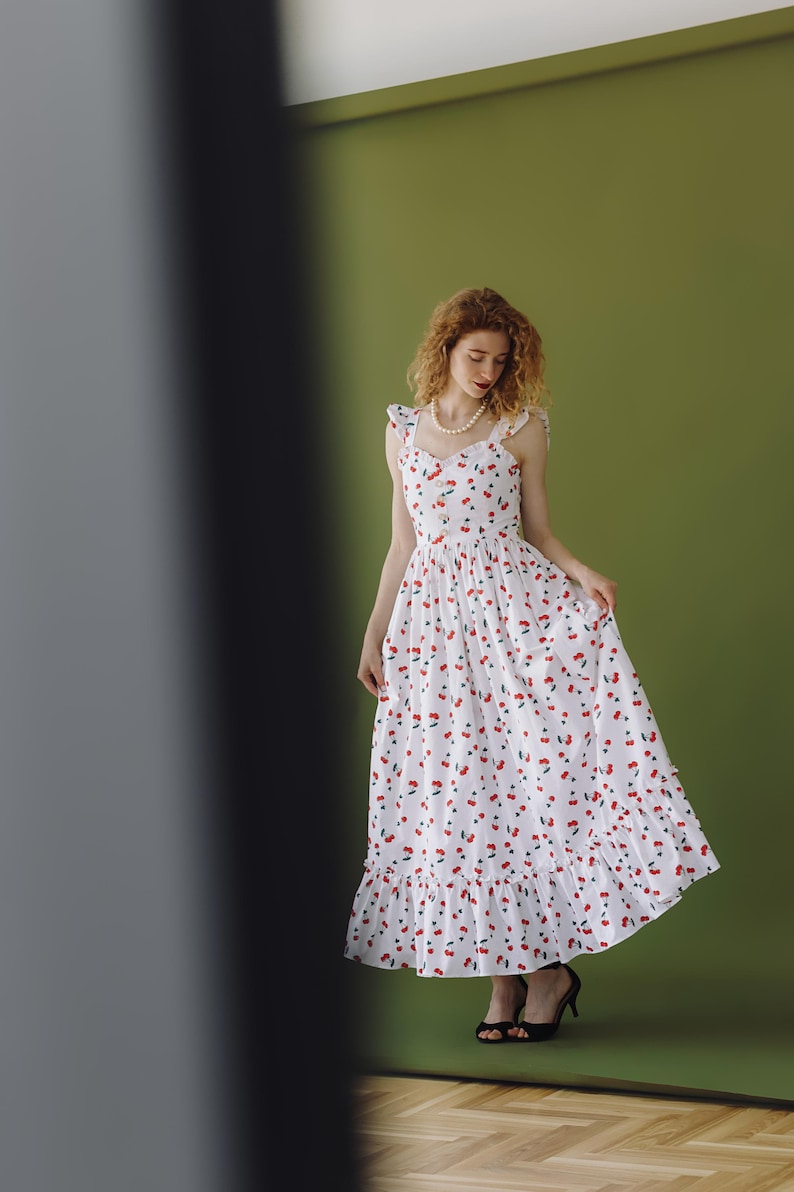 K&ouml;nnte beinhalten: Ein wei&szlig;es Maxikleid mit rotem Kirschmuster. Das &auml;rmellose Kleid hat einen R&uuml;schensaum und Tr&auml;ger. Die Person tr&auml;gt schwarze Abs&auml;tze. Der Hintergrund ist einfarbig gr&uuml;n.
