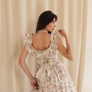 May include: A white dress featuring a pink floral pattern, a square neckline with ruffled shoulder straps, and a large fabric bow tied at the center of the back. The garment has a fitted bodice and a full skirt with a ruffled hemline detail.