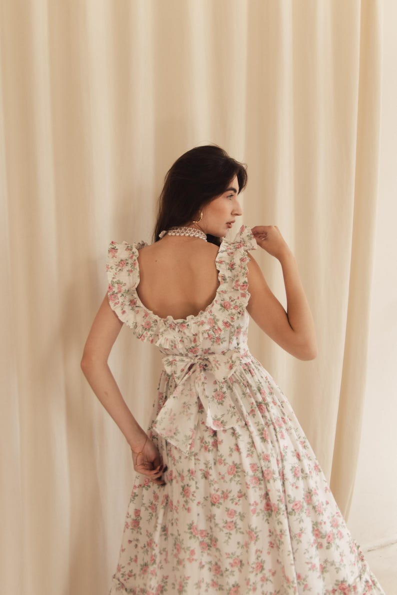May include: A white dress featuring a pink floral pattern, a square neckline with ruffled shoulder straps, and a large fabric bow tied at the center of the back. The garment has a fitted bodice and a full skirt with a ruffled hemline detail.